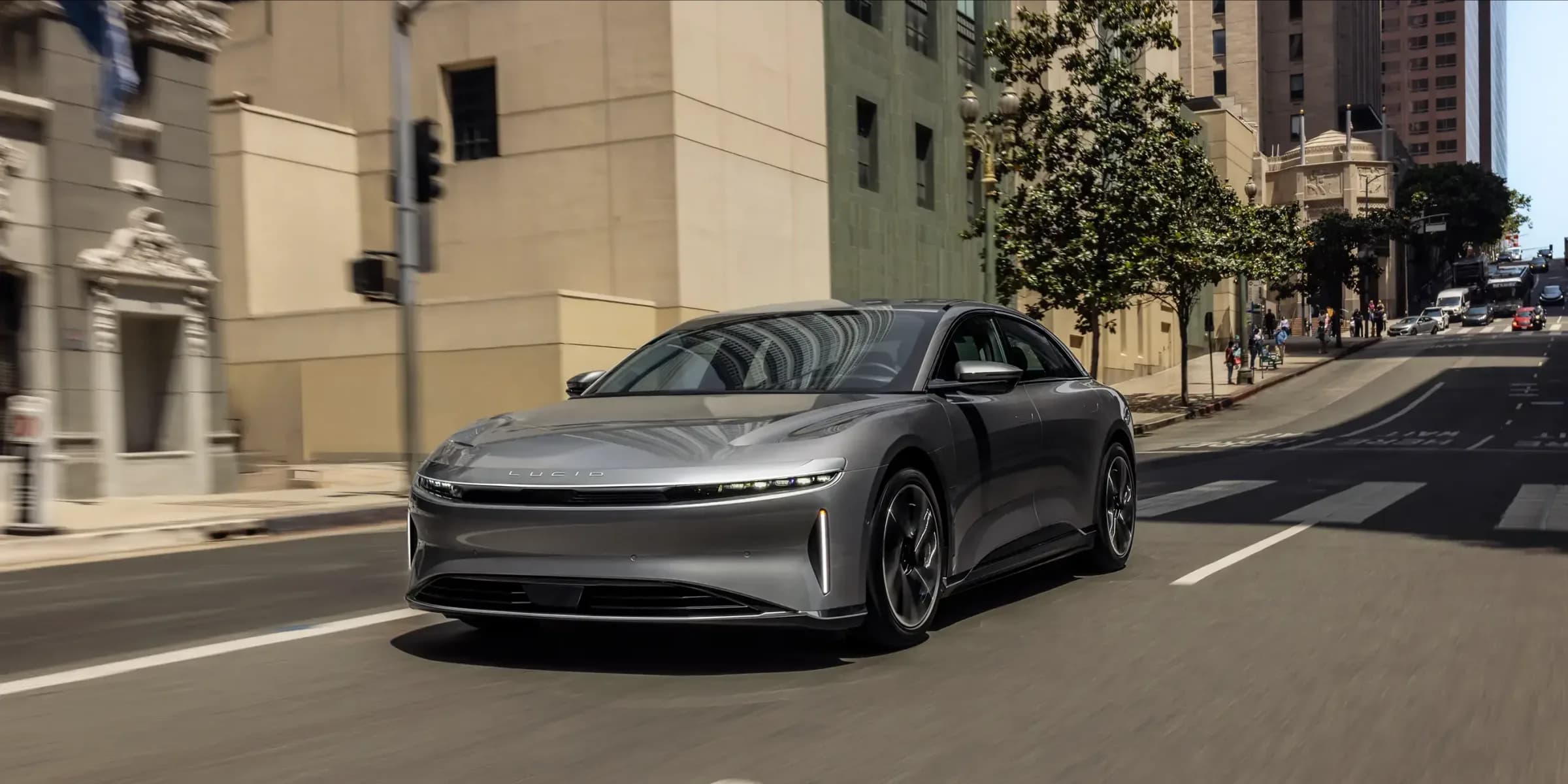 Một chiếc sedan điện Lucid Air màu bạc chạy trên con phố đô thị đầy nắng, phía sau là các toà nhà và người đi bộ.