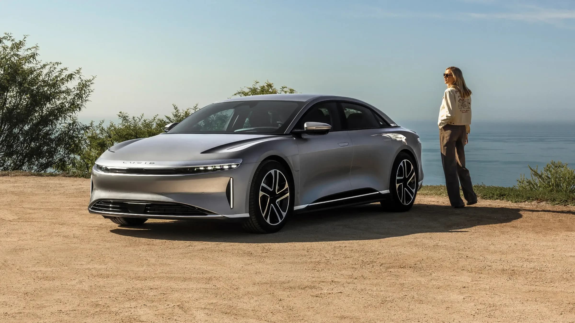 Chiếc Lucid Air màu bạc đỗ trên điểm ngắm cảnh ven biển, một người phụ nữ đứng bên cạnh.