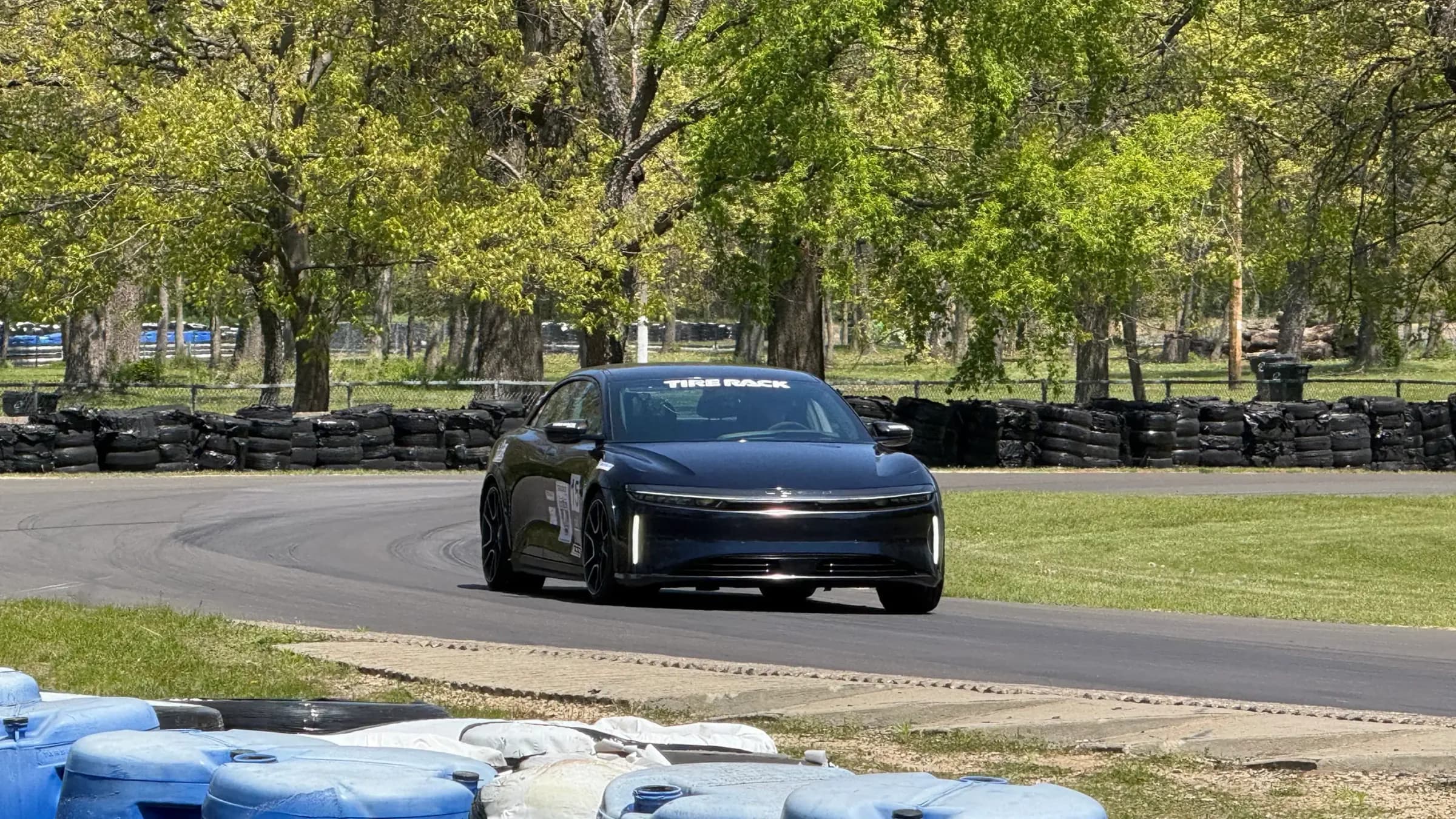 Chiếc sedan Lucid Air Sapphire vào cua trên đường đua, hai bên là tường lốp và thùng chắn màu xanh, phía sau là hàng cây.