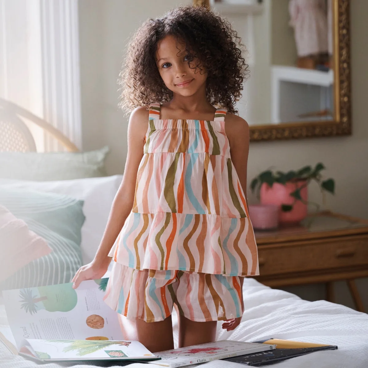Bé gái mặc pyjama kẻ sọc nhiều màu, đứng trên giường cùng sách và đồ chơi.